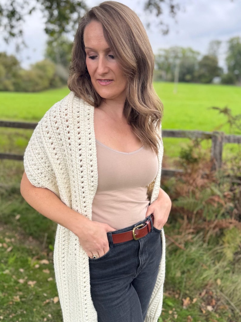 May include: Cream-colored, open-weave crochet shawl. The shawl is draped over the shoulders. The model is wearing a tan tank top, dark jeans, and a brown leather belt with a gold buckle. The background is a green field and a wooden fence.