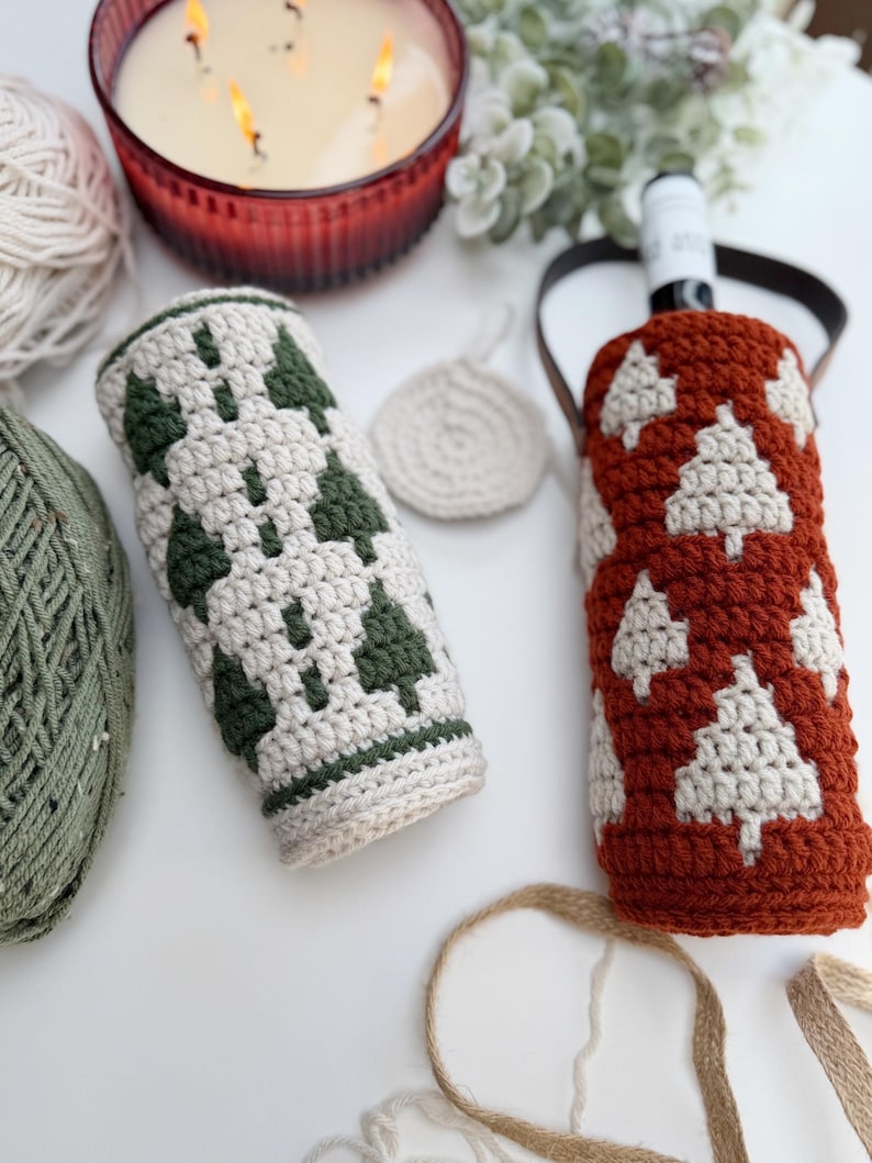 May include: Two crocheted wine bottle covers, one in cream with green tree designs, the other in rust with cream tree designs. A small cream coaster and a candle in a red glass are also visible.