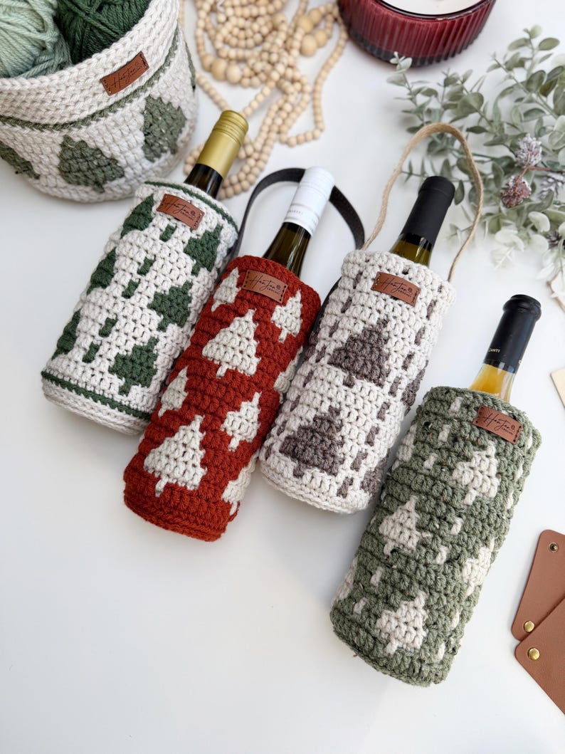 May include: Four wine bottle covers in various colors, including green, red, and gray, each with a white tree pattern. The covers are crocheted and have a leather tag. A matching basket is also visible, along with a bottle of wine.