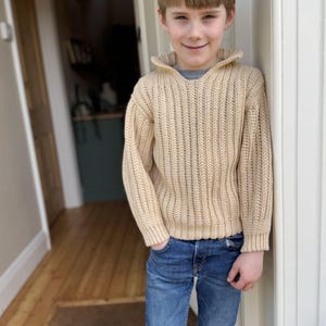 K&ouml;nnte beinhalten: Ein junges Kind tr&auml;gt einen beigefarbenen Strickpullover mit Kragen und blaue Jeans.