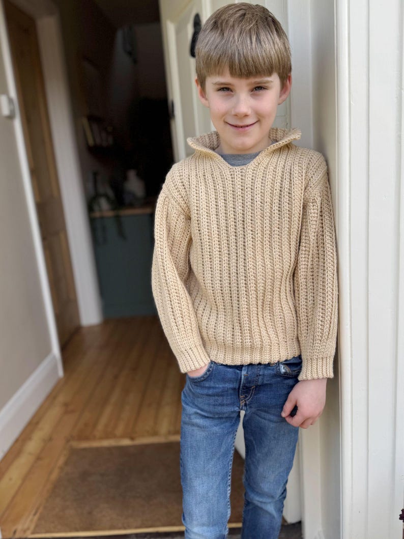 K&ouml;nnte beinhalten: Ein junges Kind tr&auml;gt einen beigefarbenen Strickpullover mit Kragen und blaue Jeans.