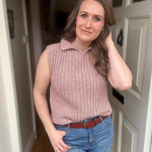 May include: A sleeveless, dusky pink knitted top with a collared neckline and a brown leather belt. The top is paired with blue jeans. The woman is standing in a doorway.