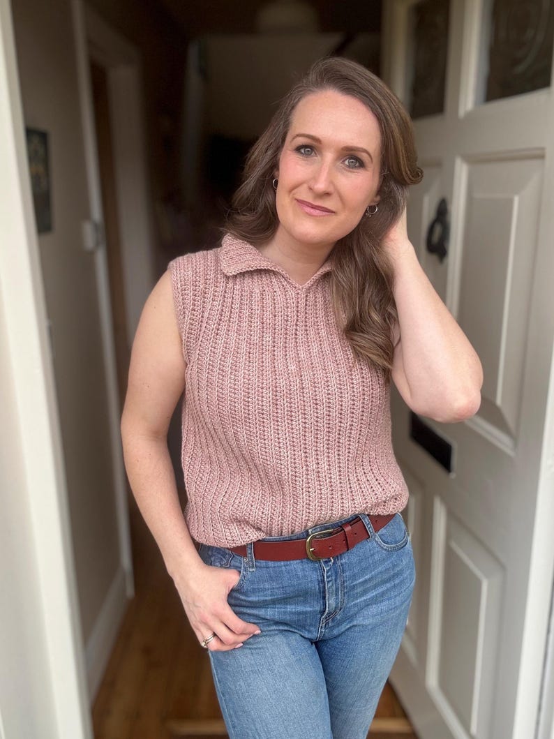 May include: A sleeveless, dusky pink knitted top with a collared neckline and a brown leather belt. The top is paired with blue jeans. The woman is standing in a doorway.