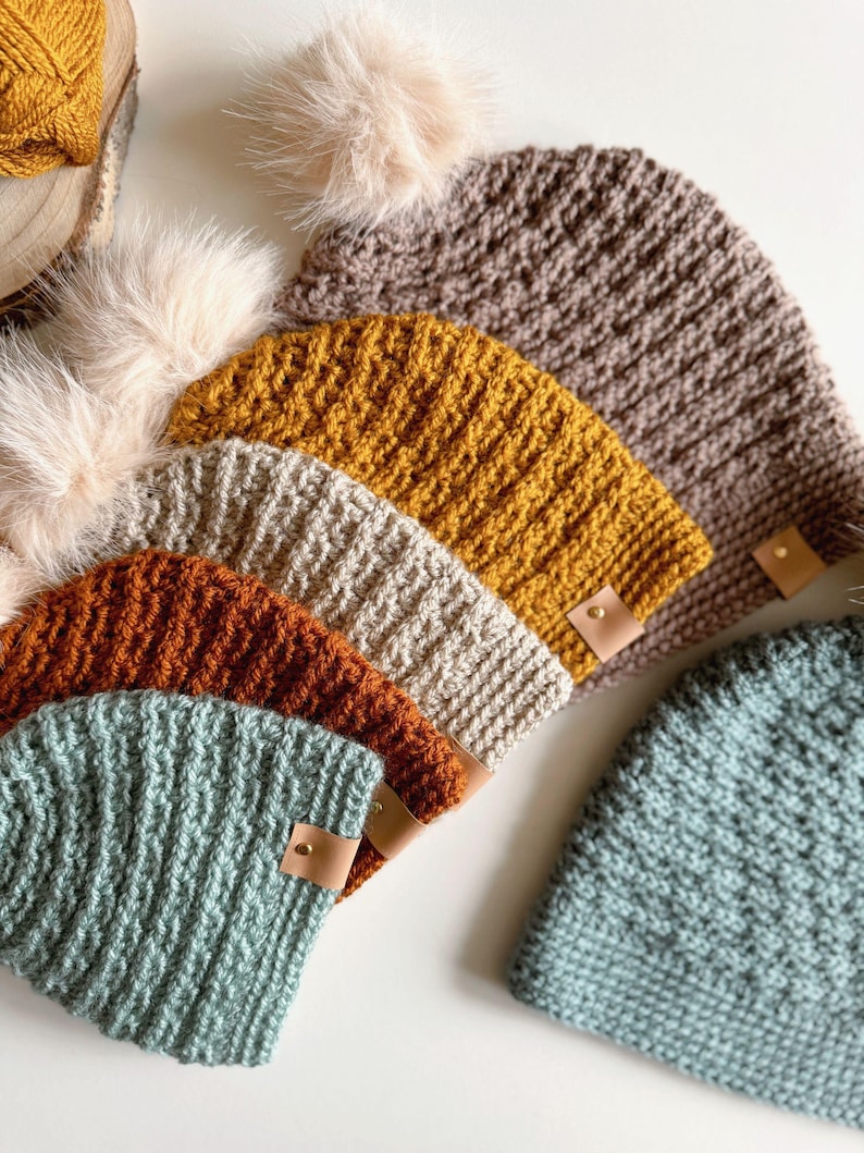 May include: A collection of handmade knit beanies in various colors, including mustard yellow, rust orange, and teal. Each beanie features a ribbed knit design and a small leather tag. Some beanies have a fluffy pom-pom.