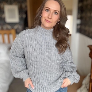 May include: A light grey, ribbed-knit jumper with a high neckline and long sleeves. The jumper is worn with blue jeans. The model is standing indoors in front of a blurred background.