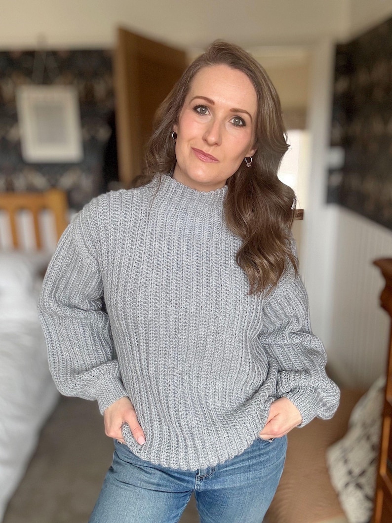 May include: A light grey, ribbed-knit jumper with a high neckline and long sleeves. The jumper is worn with blue jeans. The model is standing indoors in front of a blurred background.