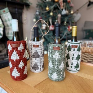 May include: Four wine bottle covers in red, cream, gray, and green, each with a crocheted Christmas tree design. The bottles are on a wooden table, with a decorated Christmas tree in the background.