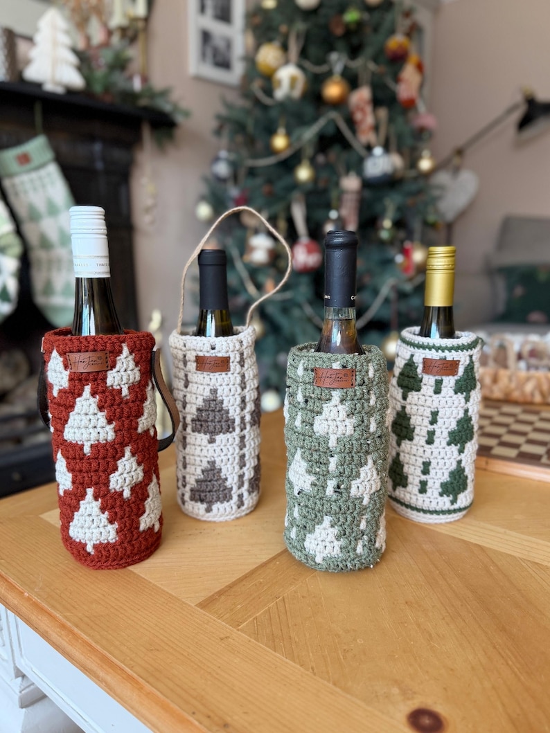 May include: Four wine bottle covers in red, cream, gray, and green, each with a crocheted Christmas tree design. The bottles are on a wooden table, with a decorated Christmas tree in the background.