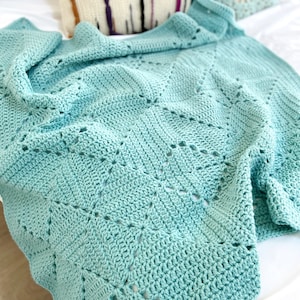 May include: A soft, crocheted throw blanket in a light teal color with a delicate, openwork pattern. The blanket is draped over a white bed with pillows.
