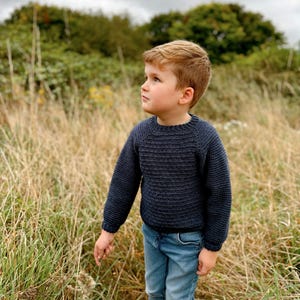 May include: A young boy wearing a dark blue knitted sweater and blue jeans stands in a field of tall grass, looking up at the sky.