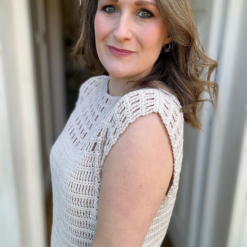 May include: A close-up of a woman wearing a beige crochet top with a delicate openwork pattern. The top has a scoop neckline and short sleeves.