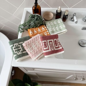 May include: A selection of six colourful, crocheted washcloths with geometric patterns. The washcloths are in shades of green, orange, pink, and burgundy, and are arranged on a white sink. The washcloths are approximately 20 cm square.