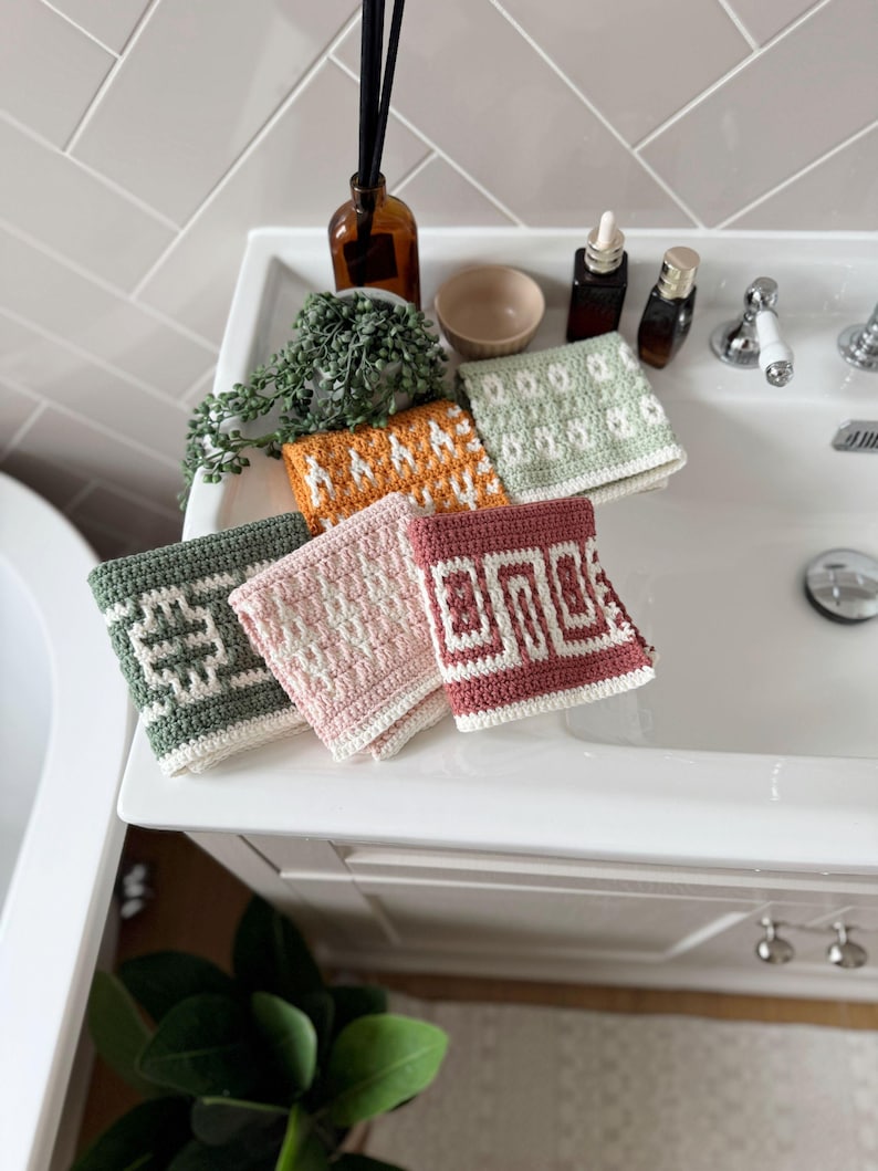 May include: A selection of six colourful, crocheted washcloths with geometric patterns. The washcloths are in shades of green, orange, pink, and burgundy, and are arranged on a white sink. The washcloths are approximately 20 cm square.
