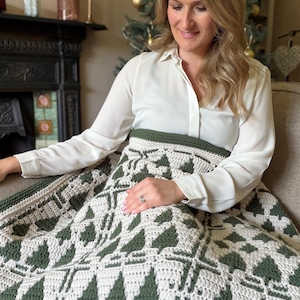 May include: A crochet blanket featuring a repeating pattern of dark forest green pine trees against a cream-colored background. The textile has a thick, textured stitch construction with a solid dark green border along the top edge.