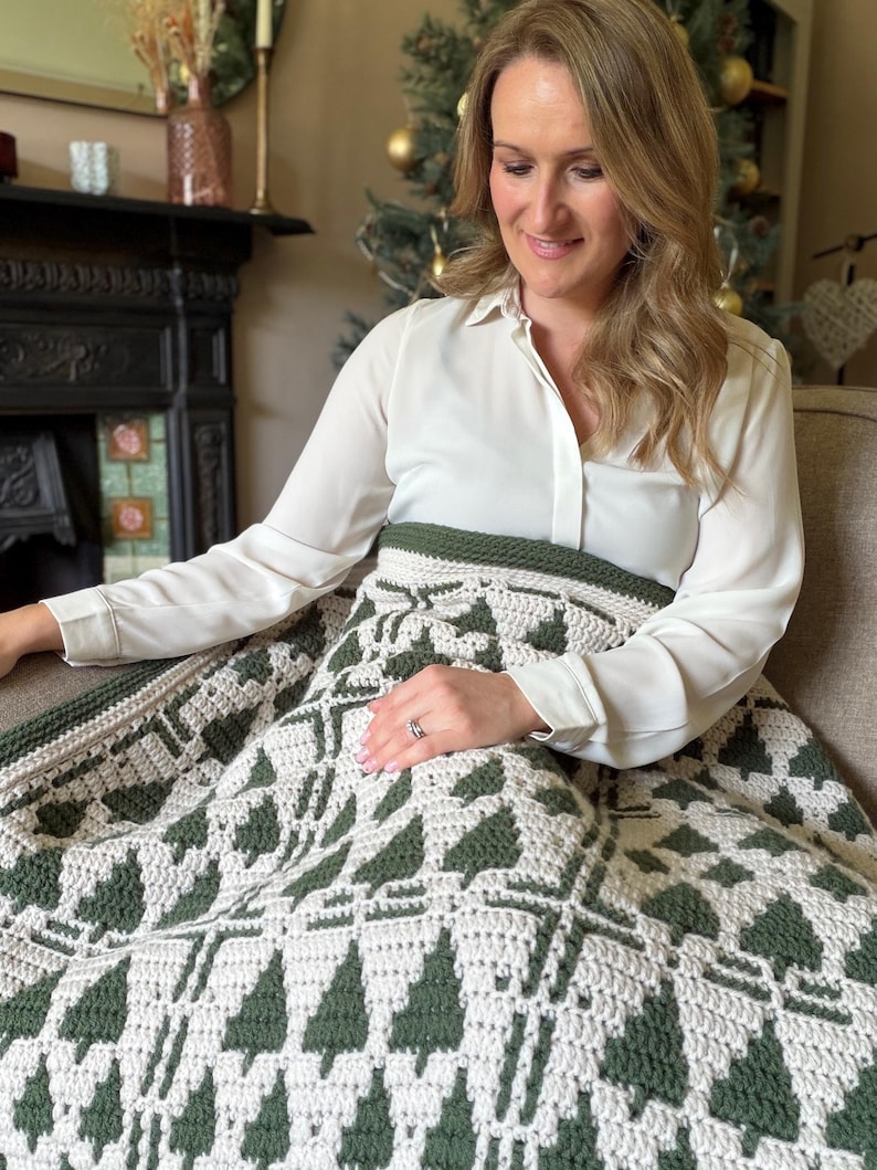 May include: A crochet blanket featuring a repeating pattern of dark forest green pine trees against a cream-colored background. The textile has a thick, textured stitch construction with a solid dark green border along the top edge.