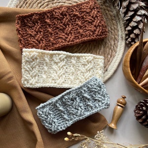 May include: Three crocheted headbands in different colors: brown, white, and gray. The headbands are laid out on a brown fabric surface with a wooden dowel and pine cones.