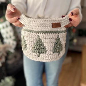 May include: A light beige crocheted basket with green Christmas tree motifs around the sides. The basket has two handles and a small brown tag. Ideal for home decor or storage, this handmade item measures approximately 20 cm in diameter.