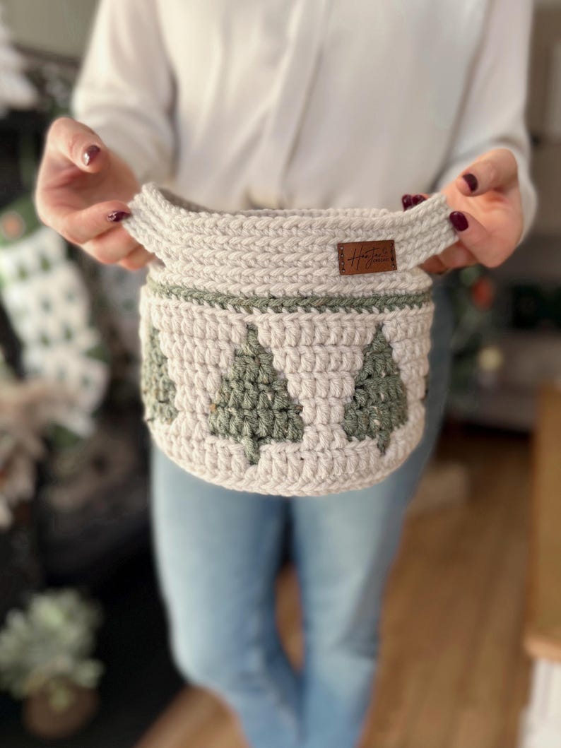 May include: A light beige crocheted basket with green Christmas tree motifs around the sides. The basket has two handles and a small brown tag. Ideal for home decor or storage, this handmade item measures approximately 20 cm in diameter.