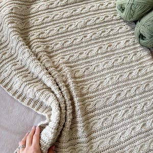 Peut inclure: Une couverture tricot&eacute;e &agrave; la main de couleur cr&egrave;me avec un motif de torsades. La couverture est pli&eacute;e, montrant la texture et le motif. Deux pelotes de fil vert sont dans le coin sup&eacute;rieur droit. La couverture est en fil doux.