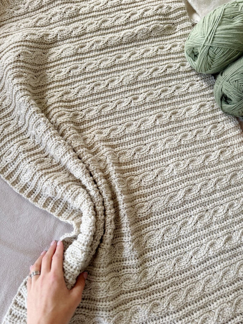 Peut inclure: Une couverture tricot&eacute;e &agrave; la main de couleur cr&egrave;me avec un motif de torsades. La couverture est pli&eacute;e, montrant la texture et le motif. Deux pelotes de fil vert sont dans le coin sup&eacute;rieur droit. La couverture est en fil doux.