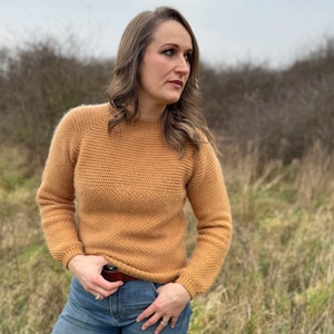 Puede incluir: Una mujer con un suéter amarillo mostaza de cuello redondo y mangas largas. Lleva pantalones vaqueros azules y un cinturón marrón.