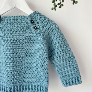 May include: A light blue crocheted sweater with a textured stitch pattern and button details on the shoulder.