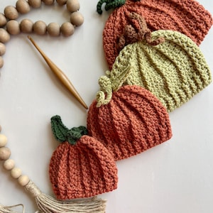 May include: Four crocheted pumpkin hats in various sizes, with green stems and brown, orange, and green yarn. The hats are arranged in a stack, with the largest hat on the bottom and the smallest hat on top.