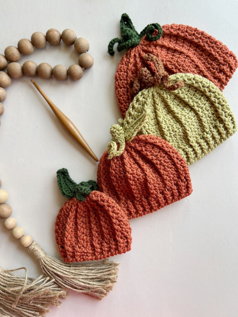 May include: Four crocheted pumpkin hats in various sizes, with green stems and brown, orange, and green yarn. The hats are arranged in a stack, with the largest hat on the bottom and the smallest hat on top.