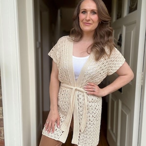 May include: Cream-colored, open-front crochet cardigan with short sleeves and a matching belt. The cardigan features a geometric pattern and is worn over a white top and brown pants. The woman is standing in a doorway.