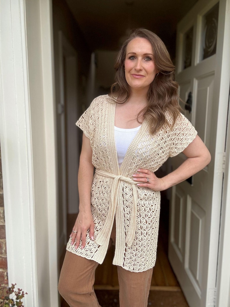 May include: Cream-colored, open-front crochet cardigan with short sleeves and a matching belt. The cardigan features a geometric pattern and is worn over a white top and brown pants. The woman is standing in a doorway.