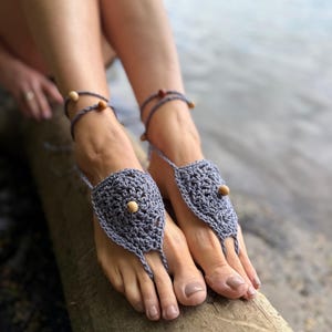 Puede incluir: Un par de sandalias descalzas de ganchillo gris con un motivo circular central y una cuenta de madera marrón claro en el centro. Las sandalias incluyen finas tiras de ganchillo que se envuelven alrededor de los tobillos con pequeñas cuentas.
