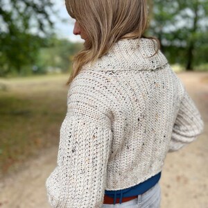 Pu&ograve; includere: Un cardigan in crochet color crema con un motivo maculato. Il cardigan ha un collo alto e maniche lunghe. Il cardigan &egrave; corto e viene indossato con un paio di jeans grigi chiari.