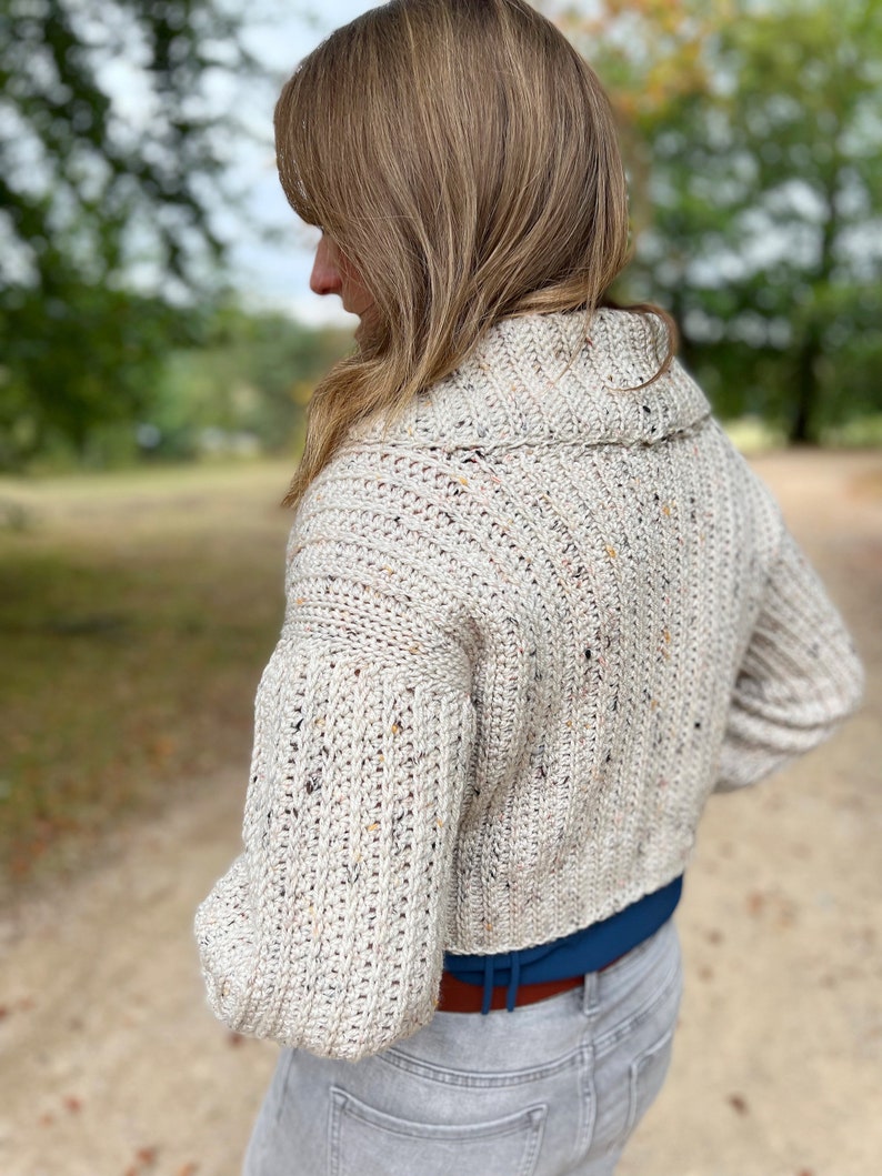 Pu&ograve; includere: Un cardigan in crochet color crema con un motivo maculato. Il cardigan ha un collo alto e maniche lunghe. Il cardigan &egrave; corto e viene indossato con un paio di jeans grigi chiari.
