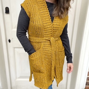 Pu&ograve; includere: Un lungo gilet di uncinetto giallo senape con scollo a V, tasche e cintura da annodare.