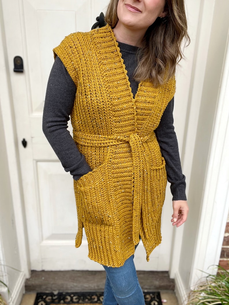 Pu&ograve; includere: Un lungo gilet di uncinetto giallo senape con scollo a V, tasche e cintura da annodare.