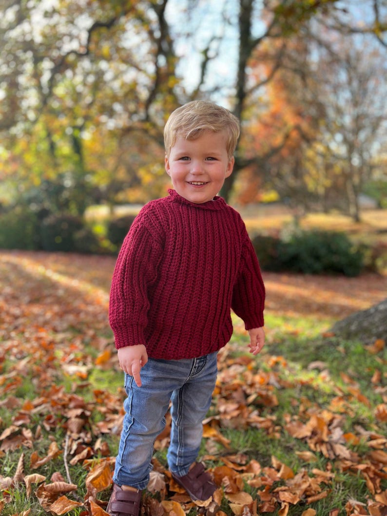 K&ouml;nnte beinhalten: Ein kleines Kind tr&auml;gt einen roten Strickpullover und eine blaue Jeans und steht in einem Haufen Herbstlaub.