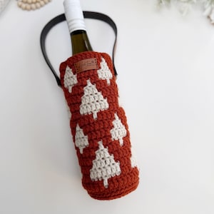 May include: A crocheted wine bottle holder in a rust-colored hue, adorned with white Christmas tree motifs. It features a black handle and a small leather tag with a handwritten signature. The bottle inside is partially visible.