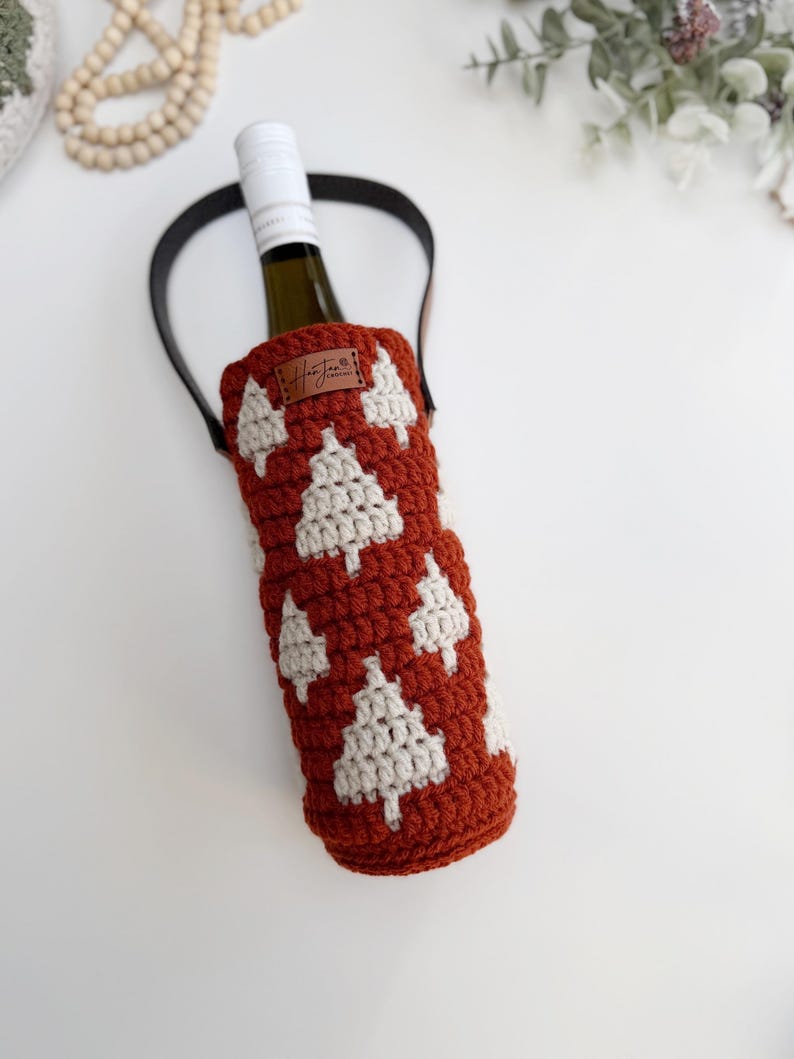 May include: A crocheted wine bottle holder in a rust-colored hue, adorned with white Christmas tree motifs. It features a black handle and a small leather tag with a handwritten signature. The bottle inside is partially visible.