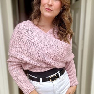 May include: A pink knitted sweater with a draped design and long sleeves. The sweater is worn over a black top and white jeans with a black belt.