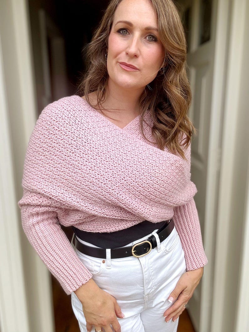 May include: A pink knitted sweater with a draped design and long sleeves. The sweater is worn over a black top and white jeans with a black belt.