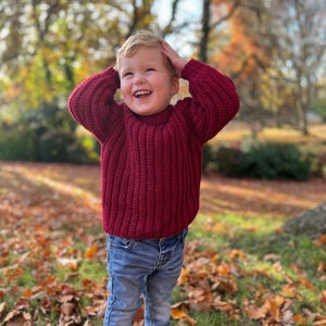 May include: A young child wearing a maroon knitted sweater and blue jeans stands in a pile of autumn leaves. The child is smiling and has their hands on their head.