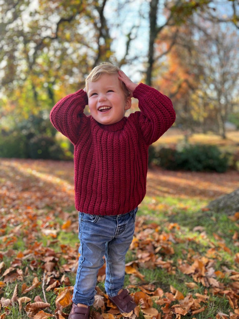 K&ouml;nnte beinhalten: Ein kleines Kind in einem weinroten Strickpullover und blauen Jeans steht in einem Haufen Herbstlaub. Das Kind l&auml;chelt und hat die H&auml;nde auf dem Kopf.