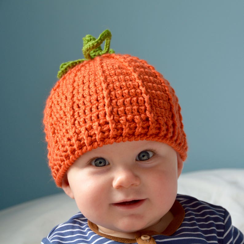 May include: A baby wearing an orange crocheted pumpkin hat with a green stem. The baby is looking at the camera and smiling.