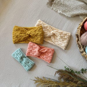May include: Four knitted headbands in various colors: mustard yellow, cream, coral, and light blue. Each headband features a twisted knot design. The headbands are arranged on a neutral-toned surface, with a basket of yarn and dried plants in the background.