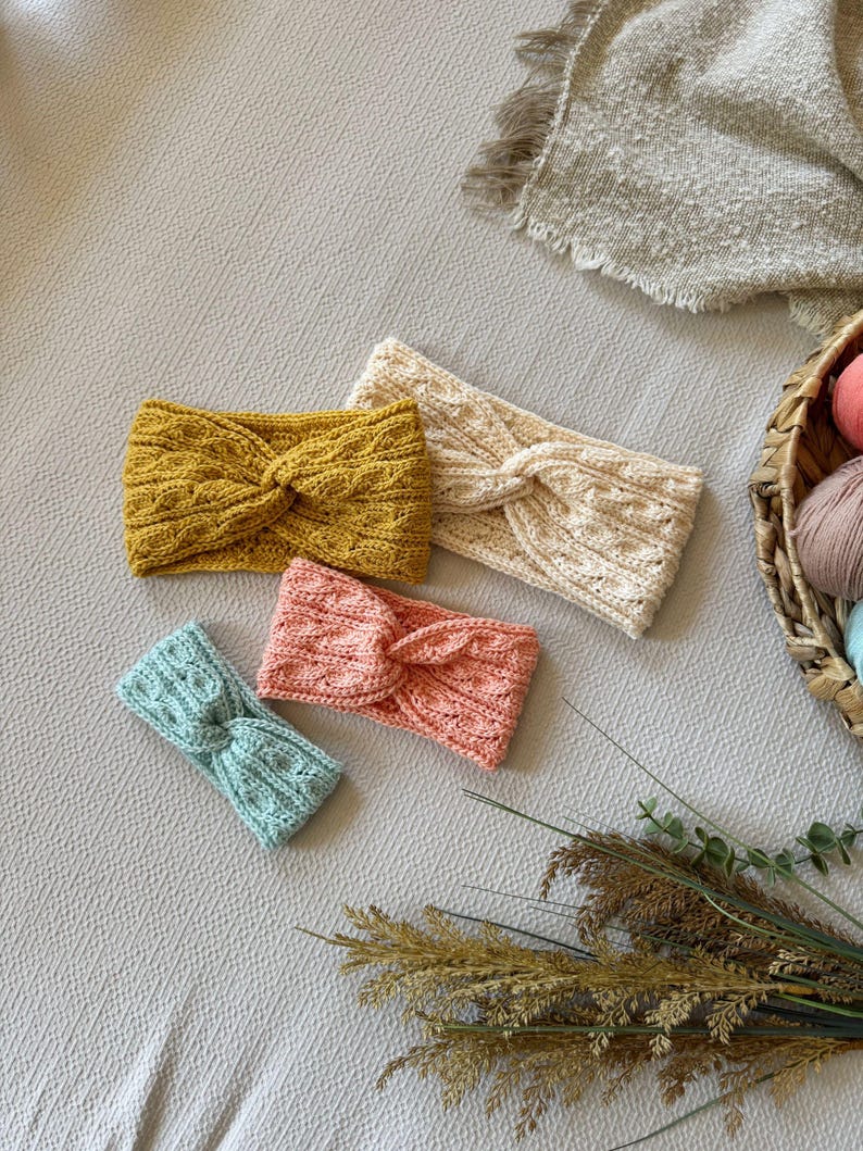 May include: Four knitted headbands in various colors: mustard yellow, cream, coral, and light blue. Each headband features a twisted knot design. The headbands are arranged on a neutral-toned surface, with a basket of yarn and dried plants in the background.