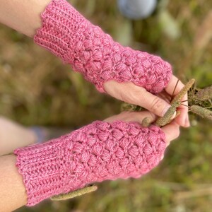 May include: A pair of pink crocheted fingerless gloves with a textured stitch pattern. The gloves are worn on a person's hands.