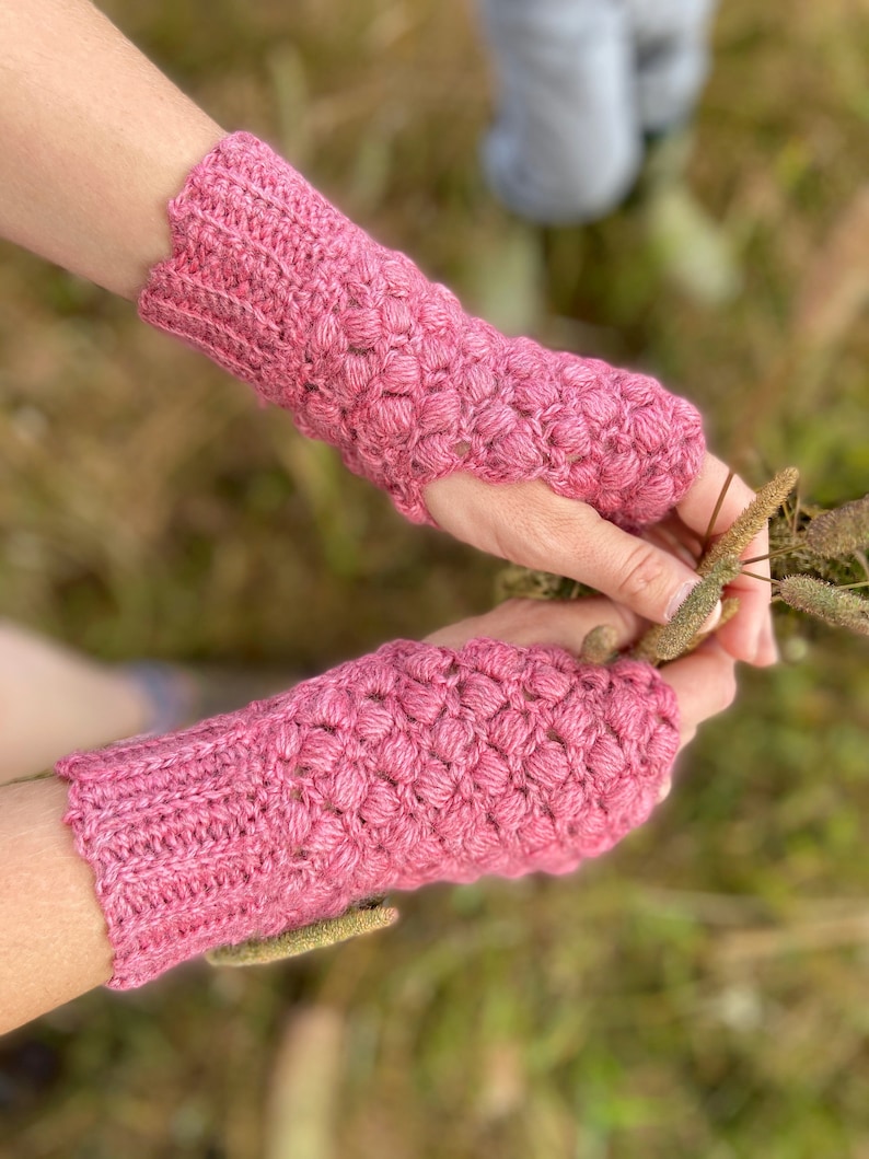 May include: A pair of pink crocheted fingerless gloves with a textured stitch pattern. The gloves are worn on a person's hands.