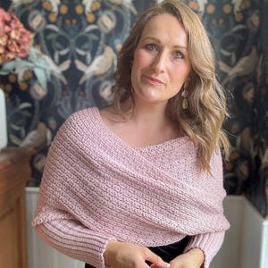 May include: A pink knitted shawl with long sleeves and a draped neckline. The shawl is worn by a person with long brown hair and is styled in a casual, relaxed way.