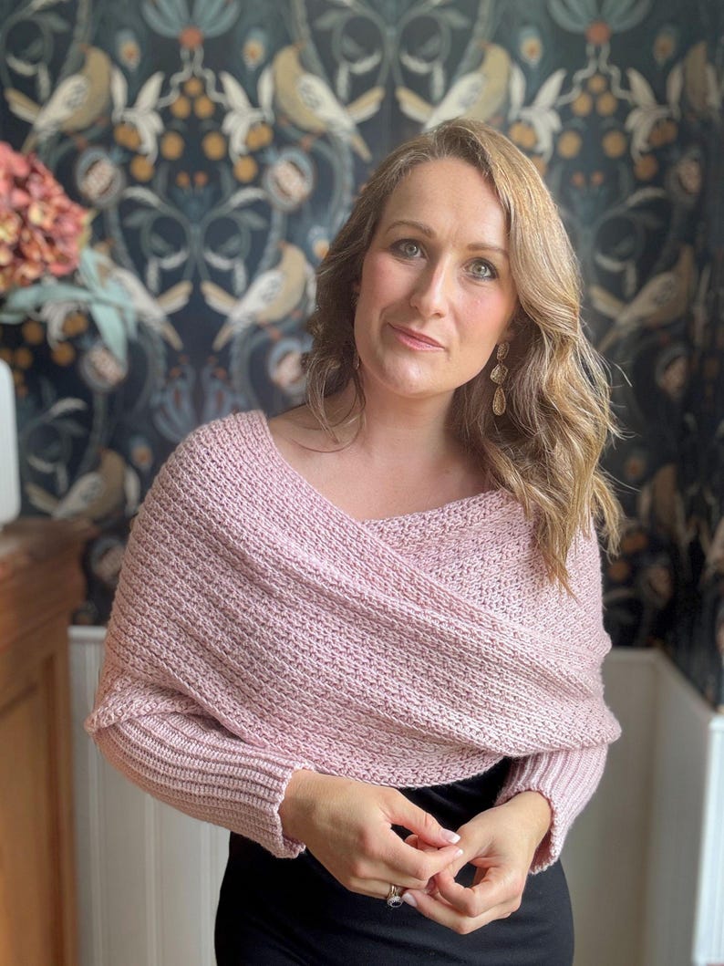 May include: A pink knitted shawl with long sleeves and a draped neckline. The shawl is worn by a person with long brown hair and is styled in a casual, relaxed way.