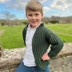 K&ouml;nnte beinhalten: Ein kleiner Junge tr&auml;gt einen dunkelgr&uuml;nen Strickcardigan mit einem wei&szlig;en Hemd mit Kn&ouml;pfen und blauen Jeans. Er steht vor einer Steinmauer mit einem grasbewachsenen Feld im Hintergrund.